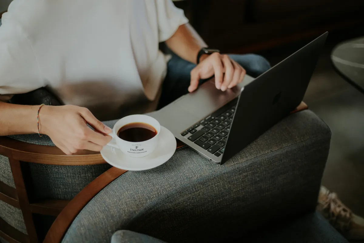 A person holding a cup of coffee while using a laptop in an armchair
